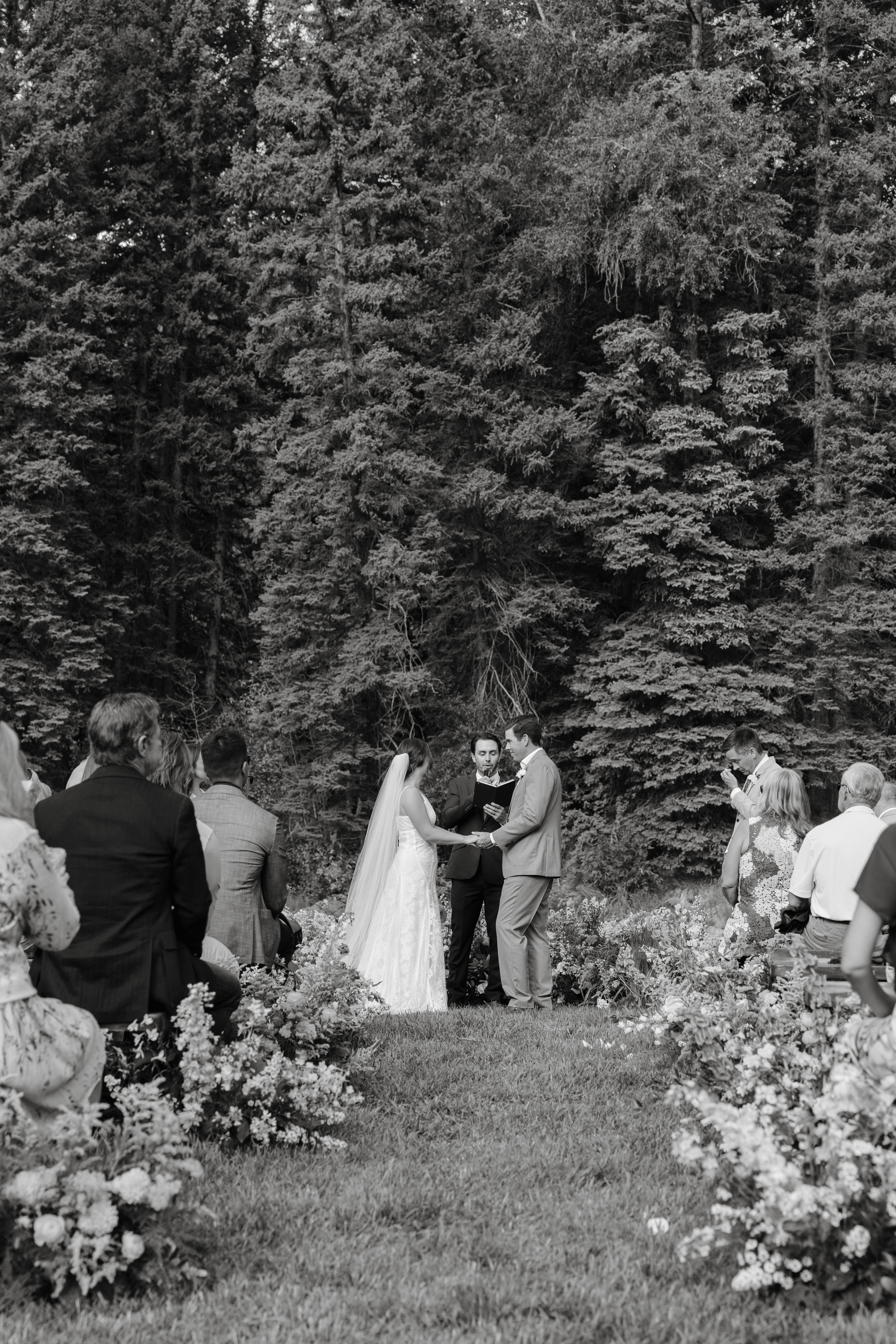 Bride and groom exchanging vows at outdoor wedding with guests and forest backdrop.